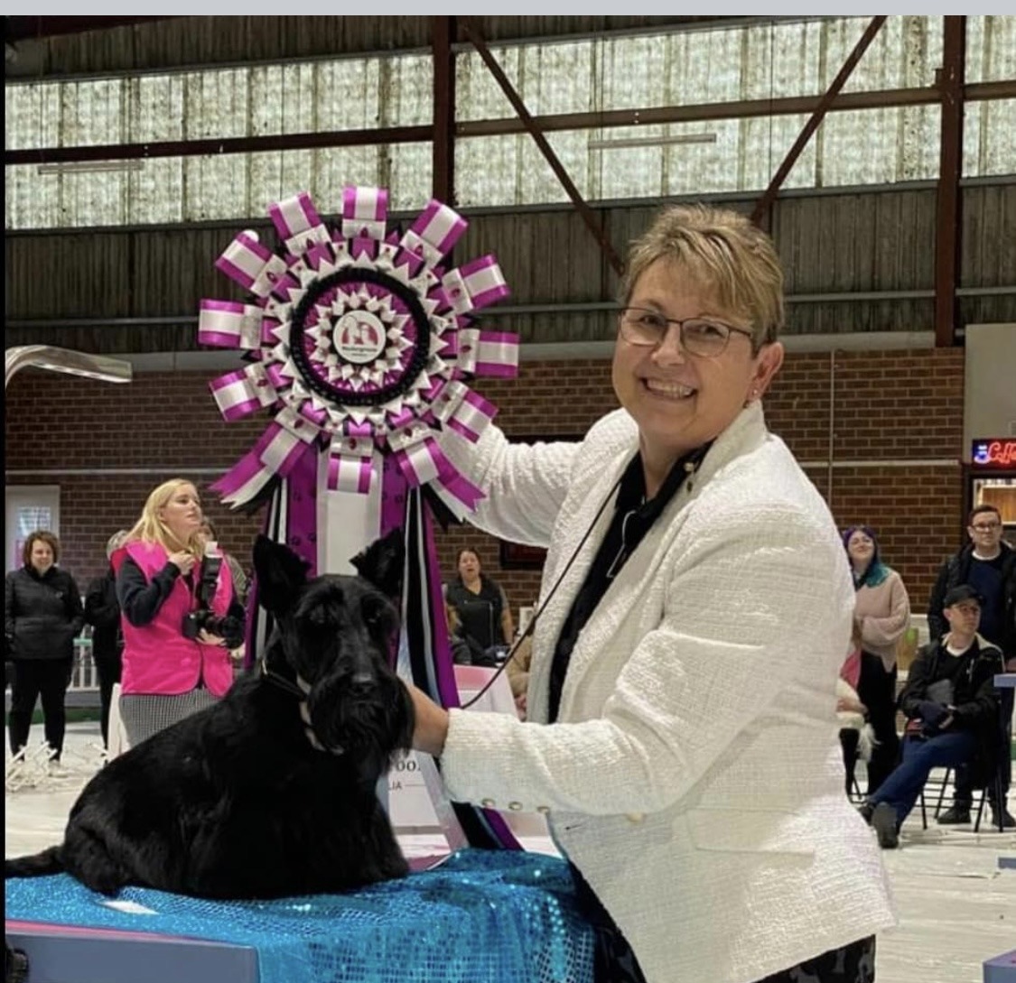 Tina in a white jacket at a dog show, standing with a black Scottish terrier beside a large show rosette